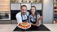 Profikoch Max Imperto und Moderatorin Tanja stehen lächelnd in einer modernen Showküche. Max präsentiert einen mit frischen Himbeeren, Erdbeeren und Schokoladensoße dekorierten Skyr-Cheesecake auf einem Teller, während Tanja eine Erdbeere in der Hand hält.