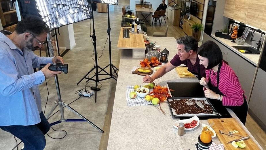 Hinter den Kulissen eines Kochvideos: Sarah und Philipp stehen in einer modernen Showroom-Küche an einer Kochinsel und betrachten ein Blech mit frisch zubereitetem Cashew-Schoko-Granola. Ein Kameramann mit professioneller Beleuchtung filmt die Szene.