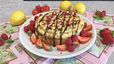 Ein gebackener Protein-Cheesecake, serviert auf einem weißen Teller und verziert mit dunkler Sauce sowie getrockneten Himbeerstückchen. Rund um den Kuchen sind frische Erdbeerscheiben und Himbeeren angerichtet, während im Hintergrund ganze Zitronen auf rosa Servietten liegen.