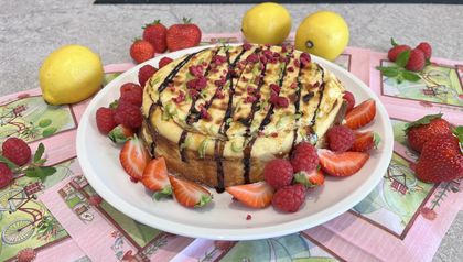 Ein gebackener Protein-Cheesecake, serviert auf einem weißen Teller und verziert mit dunkler Sauce sowie getrockneten Himbeerstückchen. Rund um den Kuchen sind frische Erdbeerscheiben und Himbeeren angerichtet, während im Hintergrund ganze Zitronen auf rosa Servietten liegen.