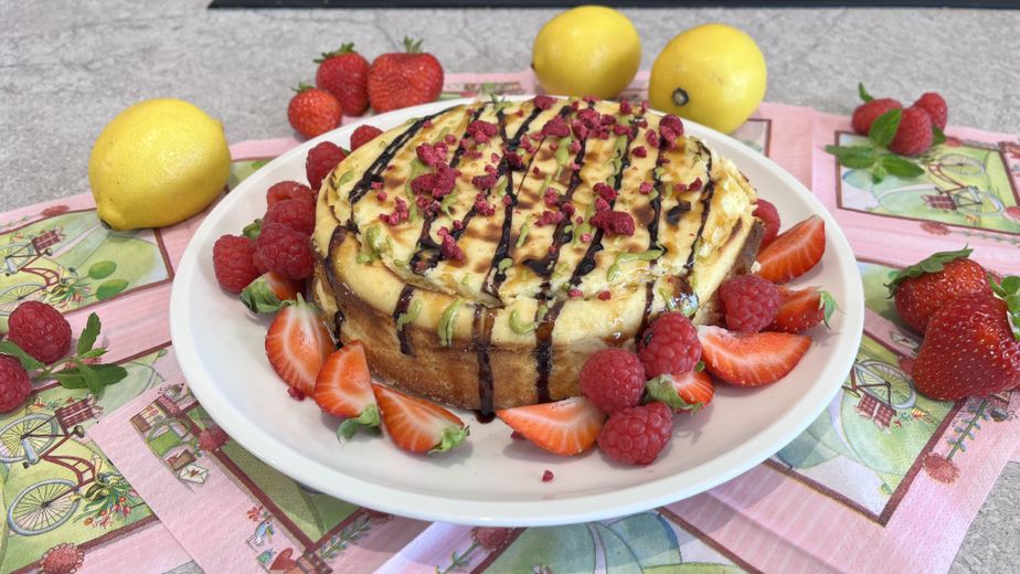 Ein gebackener Protein-Cheesecake, serviert auf einem weißen Teller und verziert mit dunkler Sauce sowie getrockneten Himbeerstückchen. Rund um den Kuchen sind frische Erdbeerscheiben und Himbeeren angerichtet, während im Hintergrund ganze Zitronen auf rosa Servietten liegen.