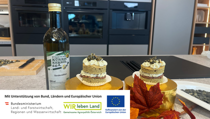 Ein fertig angerichtetes Kürbis-Törtchen, das aus zwei Schichten hellem Kuchenboden besteht. Dazwischen und obendrauf ist eine grünliche Kürbiskernöl-Creme aufgespritzt und das Törtchen ist mit gehacktem Kürbiskern-Krokant garniert.