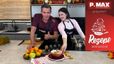 Sarah und Philipp Jelinek stehen lächelnd in einer modernen Küche und präsentieren den veganen Milchreis mit Birnenkompott. Auf der Arbeitsplatte vor ihnen sind eine fertige Portion sowie die frischen Zutaten für das Rezept angerichtet.