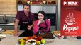 Die Moderatoren Philipp und Sarah stehen lächelnd in einer modernen Küche. Vor ihnen auf der Arbeitsplatte befindet sich ein Backblech mit dem fertigen Cashew-Schoko-Granola sowie eine angerichtete Frühstücks-Bowl mit Obst.