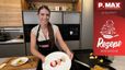 Tanja präsentiert die fertig angerichteten Topfennockerln auf einem weißen Teller mit Beerenspiegel und Puderzucker. Im Hintergrund ist eine moderne Küche mit Kochutensilien und Einbaugeräten zu sehen.