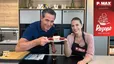 Philipp und Tanja präsentieren in einer modernen Küche ein Stück der frisch zubereiteten Schneewittchenschnitte auf einem Teller. Das Dessert zeigt deutlich seine Schichten aus hellem Teig, einer fruchtigen Himbeerfüllung, Schlagobers und einem abschließenden Butterkeks. Im Vordergrund ist das restliche Backblech mit den gleichmäßig mit Keksen belegten Schnitten zu sehen.