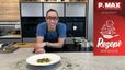 Peter Lehner präsentiert die fertig angerichteten Jakobsmuscheln mit Sauce auf einem weißen Teller, garniert mit fein gehackten Kräutern. Er steht lächelnd in einer modernen Küche hinter einer Arbeitsplatte, auf der im Hintergrund Töpfe auf dem Herd und Vorbereitungsutensilien zu sehen sind.