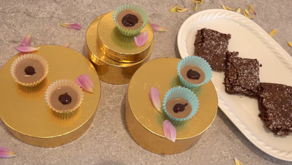 Hausgemachte Toffees in bunten Papierförmchen auf goldenen Tellern, jeweils mit einer Haselnuss und einem Schokoladenpunkt verziert, neben einem Teller mit geschnittenen veganen Kokosnuss-Brownies. Rosa Blütenblätter sind dekorativ um die Süßigkeiten verstreut.