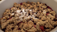 Nahaufnahme des fertigen, knusprigen Apfel-Himbeer-Crumbles in einer weißen Auflaufform. Die goldbraunen Streusel sind leicht mit Puderzucker bestäubt und bedecken die saftige rote Fruchtfüllung.