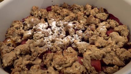 Nahaufnahme des fertigen, knusprigen Apfel-Himbeer-Crumbles in einer weißen Auflaufform. Die goldbraunen Streusel sind leicht mit Puderzucker bestäubt und bedecken die saftige rote Fruchtfüllung.