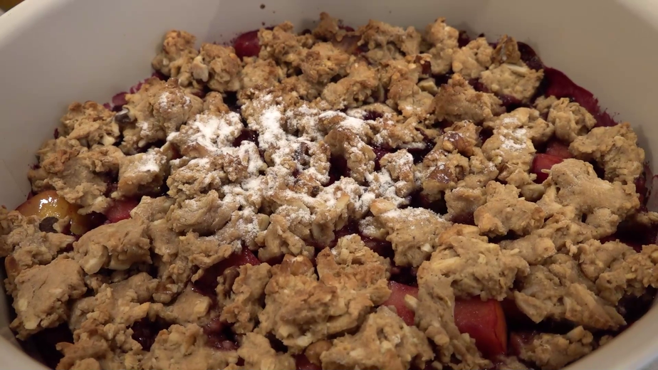 Nahaufnahme des fertigen, knusprigen Apfel-Himbeer-Crumbles in einer weißen Auflaufform. Die goldbraunen Streusel sind leicht mit Puderzucker bestäubt und bedecken die saftige rote Fruchtfüllung.