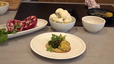 Ein fertig angerichtetes Gericht mit gebratenem Karfiol auf einem Bett aus Melanzani-Hummus, garniert mit einer Kräuter-Vinaigrette mit Granatapfelkernen. Im Hintergrund sind die frischen Zutaten zu sehen: ein aufgeschnittener Granatapfel und eine Schüssel mit Karfiolröschen.