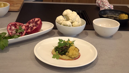 Ein fertig angerichtetes Gericht mit gebratenem Karfiol auf einem Bett aus Melanzani-Hummus, garniert mit einer Kräuter-Vinaigrette mit Granatapfelkernen. Im Hintergrund sind die frischen Zutaten zu sehen: ein aufgeschnittener Granatapfel und eine Schüssel mit Karfiolröschen.