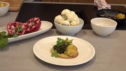 Ein fertig angerichtetes Gericht mit gebratenem Karfiol auf einem Bett aus Melanzani-Hummus, garniert mit einer Kräuter-Vinaigrette mit Granatapfelkernen. Im Hintergrund sind die frischen Zutaten zu sehen: ein aufgeschnittener Granatapfel und eine Schüssel mit Karfiolröschen.