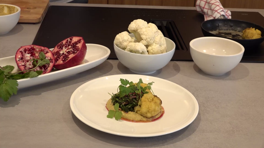 Ein fertig angerichtetes Gericht mit gebratenem Karfiol auf einem Bett aus Melanzani-Hummus, garniert mit einer Kräuter-Vinaigrette mit Granatapfelkernen. Im Hintergrund sind die frischen Zutaten zu sehen: ein aufgeschnittener Granatapfel und eine Schüssel mit Karfiolröschen.