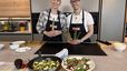 Sängerin Virginia Ernst und Peter Lehner stehen lächelnd in einer modernen Küche und halten frischen grünen Spargel in den Händen. Im Vordergrund sind die fertig zubereiteten Gerichte auf einem Holzbrett angerichtet: eine Spargel-Frittata direkt in der Pfanne sowie ein bunter Spargelsalat mit in Schinken gewickelten Stangen in einer weißen Schüssel.