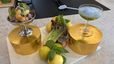Ein hellgrüner Basilikum-Cocktail und ein Garnelensalat mit lila Blüten, serviert in eisgekühlten Gläsern auf runden, goldenen Podesten. Zwischen den Gerichten sind frische Zutaten wie Zitronen, Basilikum und Römersalatherzen dekorativ auf einer hellen Küchenarbeitsplatte angerichtet.