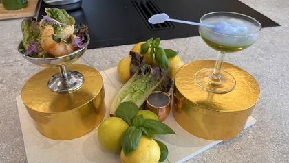 Ein hellgrüner Basilikum-Cocktail und ein Garnelensalat mit lila Blüten, serviert in eisgekühlten Gläsern auf runden, goldenen Podesten. Zwischen den Gerichten sind frische Zutaten wie Zitronen, Basilikum und Römersalatherzen dekorativ auf einer hellen Küchenarbeitsplatte angerichtet.