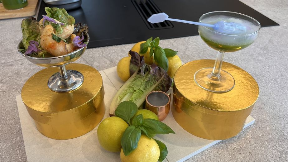 Ein hellgrüner Basilikum-Cocktail und ein Garnelensalat mit lila Blüten, serviert in eisgekühlten Gläsern auf runden, goldenen Podesten. Zwischen den Gerichten sind frische Zutaten wie Zitronen, Basilikum und Römersalatherzen dekorativ auf einer hellen Küchenarbeitsplatte angerichtet.