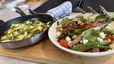 Eine Pfanne mit einer goldgelben Spargel-Frittata, verfeinert mit Spinat und Feta, steht neben einer Schüssel mit einem bunten Spargelsalat. Der Salat ist mit in Schinken gewickelten Spargelstangen, Avocado, Kirschtomaten und Feta-Bröseln angerichtet.