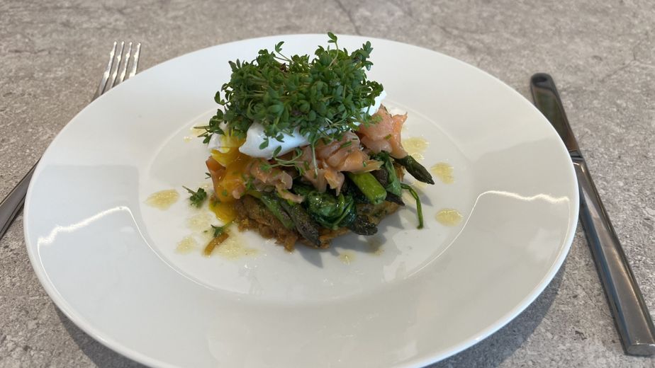 Ein knuspriger Rösti, belegt mit grünem Spargel, Räucherlachs und einem pochierten Ei. Das Gericht ist mit frischer Kresse garniert und auf einem weißen Teller angerichtet.