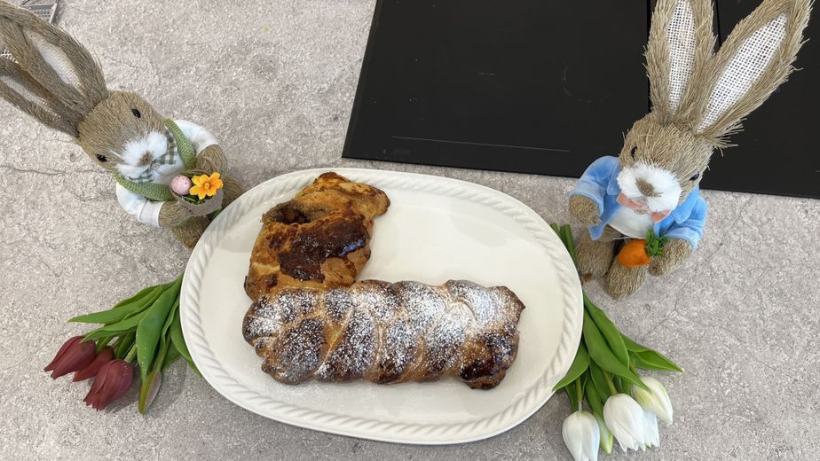 Ein fertig gebackener, goldbrauner Osterzopf in Hasenform auf einer weißen Servierplatte, der fein mit Puderzucker bestäubt ist. Das Gebäck ist festlich angerichtet zwischen zwei dekorativen Stroh-Osterhasen sowie roten und weißen Tulpen auf einer Küchenarbeitsplatte.