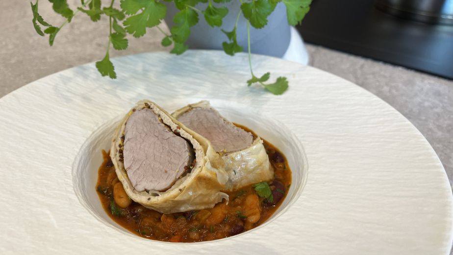 Zwei Scheiben Schweinsfilet im goldbraunen Strudelteig, angerichtet auf einem herzhaften Bohnen-Cassoulet mit weißen und Kidneybohnen. Das Gericht wird in einer weißen, strukturierten Schüssel serviert, im Hintergrund ist eine grüne Kräuterpflanze erkennbar.