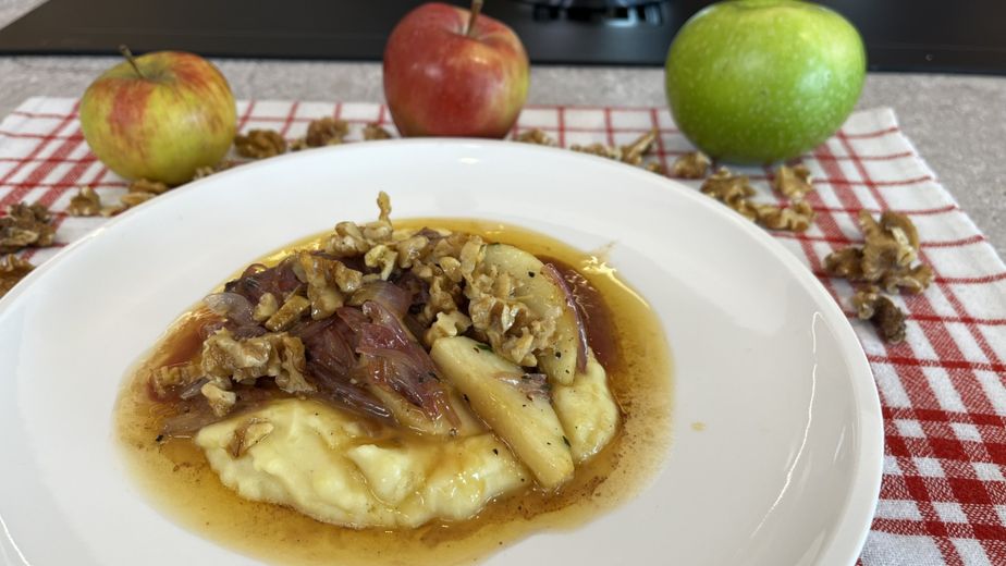 Das fertig angerichtete Gericht auf einem weißen Teller: Auf einem Bett aus cremigem Erdäpfel-Stampf sind glasierte Apfelspalten und rosa gebratene Kalbsleber in einer Sauce angerichtet und mit gehackten Walnüssen bestreut. Im Hintergrund sind als Dekoration ganze Äpfel und Walnüsse zu sehen.