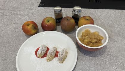 Drei mit Staubzucker bestreute Topfennockerln auf einem roten Fruchtspiegel, serviert neben einer Schüssel mit stückigem Apfelmus. Im Hintergrund ergänzen vier frische Äpfel und Gewürzdosen wie Zimt das fertig angerichtete Dessert auf einer grauen Küchenarbeitsplatte.