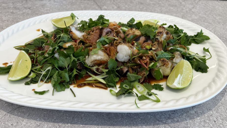 Servierfertiges Moo Nam Tok auf einer weißen ovalen Platte, garniert mit reichlich frischem Koriander, Minze, Frühlingszwiebeln und Limettenspalten. Die gegrillten Schweinefleischstreifen sind mit einer dunklen Sauce und gemörsertem Reis vermengt.