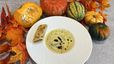 Eine weiße Schale mit der samtigen Kokos-Kürbissuppe, garniert mit dunklen Öltropfen. Am Tellerrand liegt ein Stück des fertigen Kürbisstrudels, umgeben von einer herbstlichen Dekoration aus bunten Blättern und verschiedenen Kürbissen.