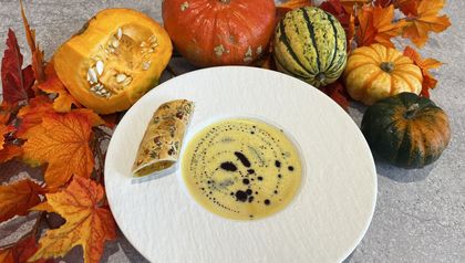 Eine weiße Schale mit der samtigen Kokos-Kürbissuppe, garniert mit dunklen Öltropfen. Am Tellerrand liegt ein Stück des fertigen Kürbisstrudels, umgeben von einer herbstlichen Dekoration aus bunten Blättern und verschiedenen Kürbissen.