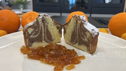 Zwei Hälften eines kleinen, mit Puderzucker bestäubten Marmorkuchens zeigen die feine hell-dunkle Teigmusterung auf einem weißen Teller. Im Vordergrund ist eine Portion glänzendes Kumquats-Kompott angerichtet, während im Hintergrund frische Orangen und Kumquats die fruchtigen Zutaten des Rezepts unterstreichen.