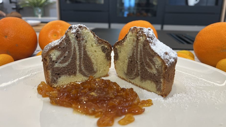 Zwei Hälften eines kleinen, mit Puderzucker bestäubten Marmorkuchens zeigen die feine hell-dunkle Teigmusterung auf einem weißen Teller. Im Vordergrund ist eine Portion glänzendes Kumquats-Kompott angerichtet, während im Hintergrund frische Orangen und Kumquats die fruchtigen Zutaten des Rezepts unterstreichen.