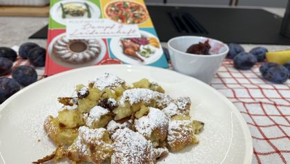 Ein Teller mit frisch zubereitetem, fluffigem Kaiserschmarrn, der großzügig mit Staubzucker bestreut ist. Im Hintergrund stehen eine kleine Schale mit Zwetschkenröster, frische Zwetschken und das Kochbuch „Kochen mit Dampf & Leidenschaft“ auf einem rot-weiß karierten Küchentuch.