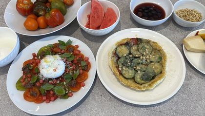 Die zwei auf der Seite beschriebenen Gerichte, angerichtet auf einem grauen Tisch: links ein bunter Tomatensalat mit Burrata, Wassermelone und Olivenpesto, rechts eine runde Tarte Tatin mit Zucchinischeiben. Im Hintergrund stehen Schalen mit frischen Zutaten wie Paradeiser-Raritäten, Pinienkernen und Oliven.