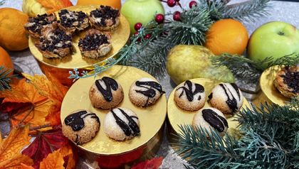 Die fertig gebackenen veganen Florentiner und Kokosmakronen sind auf goldenen Podesten angerichtet. Die runden Kokosbällchen und die flachen, nussigen Florentiner sind jeweils mit dunklen Schokoladenstreifen verziert. Das Gebäck ist festlich zwischen Tannenzweigen, Äpfeln und Orangen dekoriert.