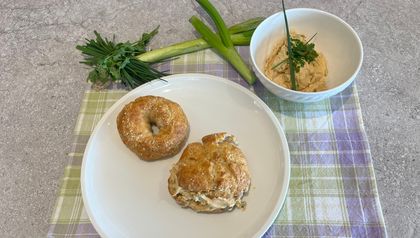 Zwei goldbraun gebackene Skyr-Bagels auf einem weißen Teller, wobei einer aufgeschnitten und reichlich mit cremigen Liptauer-Aufstrich gefüllt ist. Daneben steht eine Schüssel mit dem orangefarbenen Aufstrich, garniert mit frischen Kräutern, umgeben von Zutaten wie Frühlingszwiebeln und Schnittlauch auf einem karierten Küchentuch.