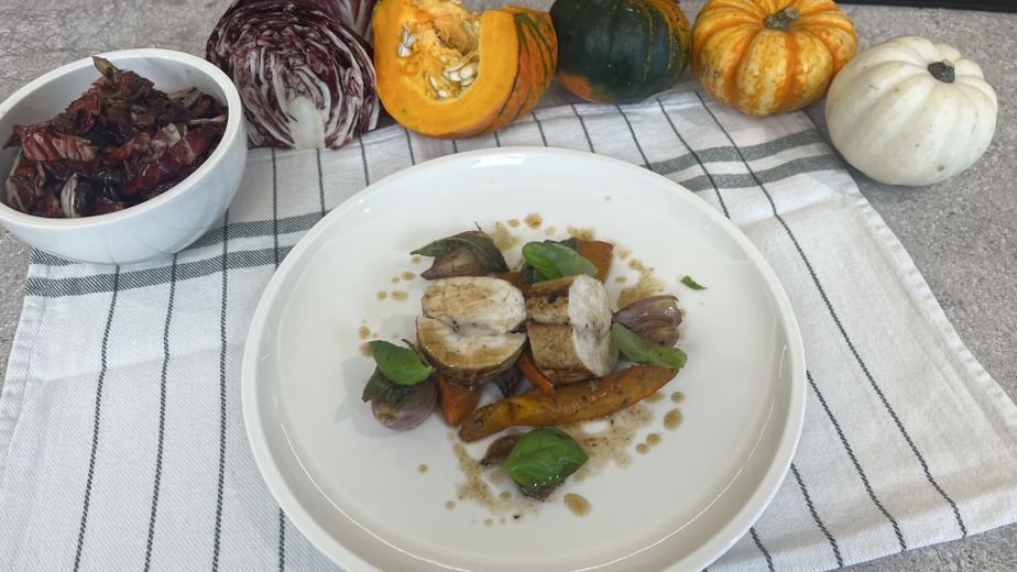 Servierfertig angerichtete Hühner-Roulade in Scheiben auf gebratenem Kürbisgemüse und Schalotten, garniert mit Basilikum. Daneben steht eine Schüssel mit dem marinierten Radicchio-Salat, während im Hintergrund die frischen Rohzutaten wie verschiedene Kürbisse und Radicchio arrangiert sind.