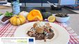 Ein weißer Teller mit knusprigen Käferbohnen-Kroketten auf dunkler Kernöl-Mayonnaise, garniert mit frisch geriebenem Kren und knusprigen Speckwürfeln. Im Hintergrund stehen eine Flasche Steirisches Kürbiskernöl, verschiedene Kürbisse, eine Schale mit Käferbohnen und geröstete Kürbiskerne auf einem rot-weiß karierten Tuch.