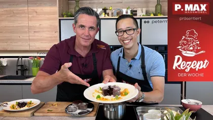 Philipp und Koch Peter Lehner stehen lächelnd in einer Küche und präsentieren das fertige Gericht auf einem Teller. Zu sehen ist der aufgeschnittene Schokokuchen mit flüssigem Kern, garniert mit Ananas-Karamell-Sauce und Mascarpone-Creme.