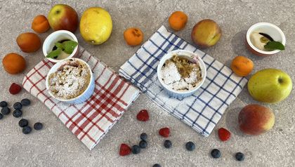 Zwei fertige Früchte Crumbles in weißen Förmchen, mit Puderzucker bestäubt. Sie sind auf einer Arbeitsplatte angerichtet, umgeben von den frischen Zutaten des Rezepts wie Äpfeln, Pfirsichen, Marillen und Beeren.