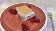 Eine servierfertige Schneewittchenschnitte auf einem roten Teller mit konzentrischen Kreisen, umgeben von einigen Himbeeren. Das quadratische Kuchenstück zeigt deutlich die Schichten aus hellem Boden, fruchtiger Himbeerfüllung und Schlagobers, gekrönt von einem ganzen Butterkeks.