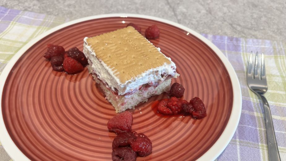 Eine servierfertige Schneewittchenschnitte auf einem roten Teller mit konzentrischen Kreisen, umgeben von einigen Himbeeren. Das quadratische Kuchenstück zeigt deutlich die Schichten aus hellem Boden, fruchtiger Himbeerfüllung und Schlagobers, gekrönt von einem ganzen Butterkeks.