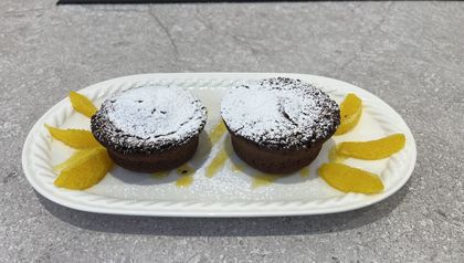 Zwei kleine Schokoküchlein, bestäubt mit Puderzucker, serviert auf einem weißen ovalen Teller. Die Küchlein sind mit frischen Orangenspalten und einem glänzenden Spiegel aus Orangensauce ansprechend garniert.