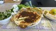 Eine frisch gebackene, goldbraun krustige Spinat-Lauch-Quiche auf Backpapier, bei der ein Anschnitt die cremige Füllung zeigt. Um die Quiche herum sind Zutaten wie Lauch, Zitronen und Parmesan sowie eine Schüssel Blattsalat und österliche Dekoration mit weißen Tulpen und einem Osterei auf einer Tischdecke arrangiert.
