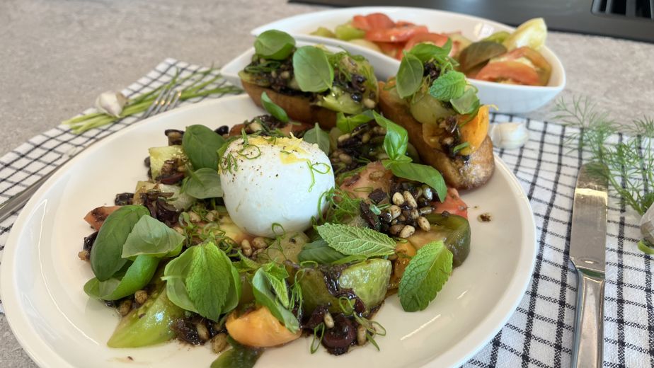 Ein weißer Teller mit einer Kugel Mozzarella in der Mitte, garniert mit Zitronenabrieb und frischen Kräutern. Um den Käse herum sind bunte Paradeiser-Stücke mit einem dunklen Oliven-Dressing, gerösteten Pinienkernen, Basilikum und Minze angerichtet. Im Hintergrund ergänzen zwei goldbraun geröstete Bruschetta-Scheiben, die mit der gleichen Tomaten-Oliven-Mischung belegt sind, das sommerliche Gericht.