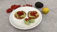 Zwei goldbraun gebackene Cloud-Bread-Fladen auf einem weißen Teller, großzügig belegt mit einer Avocado-Hüttenkäse-Mischung und gewürfelten Kirschtomaten. Im Hintergrund sind eine ganze Avocado, eine Zitrone und lose Kirschtomaten als Zutaten auf einer grauen Arbeitsfläche angerichtet.