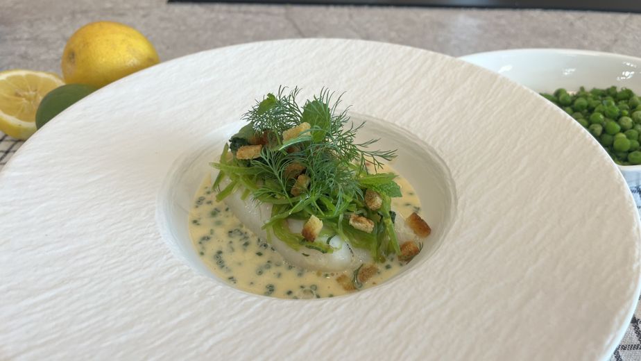 Ein tiefes, strukturiertes weißes Teller präsentiert den fertig angerichteten Kabeljau. Das Fischfilet ruht in einer cremigen Riesling Beurre Blanc, die mit feinem Schnittlauch und Forellenkaviar durchzogen ist. Getoppt wird das Gericht mit einem leuchtend grünen Zuckerschotensalat, frischen Kräutern wie Dill und Minze sowie knusprigen Croutons.