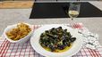 Ein fertig angerichteter Teller Moules Frites in einer hellen Sauce mit Kräutern und Gemüse, serviert mit einer Schüssel goldbraun frittierter Pommes Frites und einem Glas Weißwein. Das Ensemble ist auf einem rot-weiß karierten Tuch in einer modernen Küche vor einem schwarzen Induktionskochfeld präsentiert.